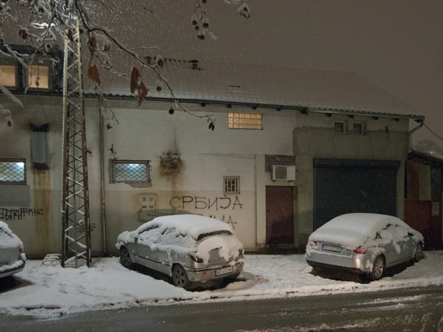 A narrow street after snowfall. Parked cars occupy the roadside space, marking the pressure of private motorization on an already compressed urban fabric.