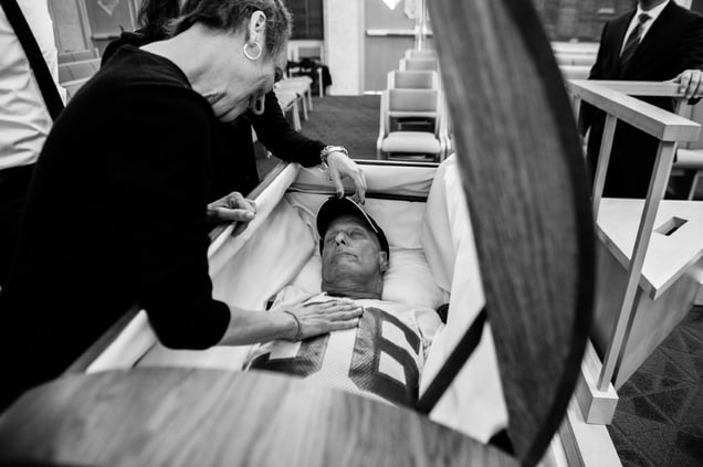 Howard Borowick left instructions for his funeral because he was a planner, and liked to be in charge, even in death. He requested to be buried in his favorite Giant’s football jersey, a pair of jeans, and his HB baseball cap. Laurel Borowick, his wife, smiles at him as they spend their last moment together before the casket is closed and the funeral begins. Mt. Kisco, New York. December 2013