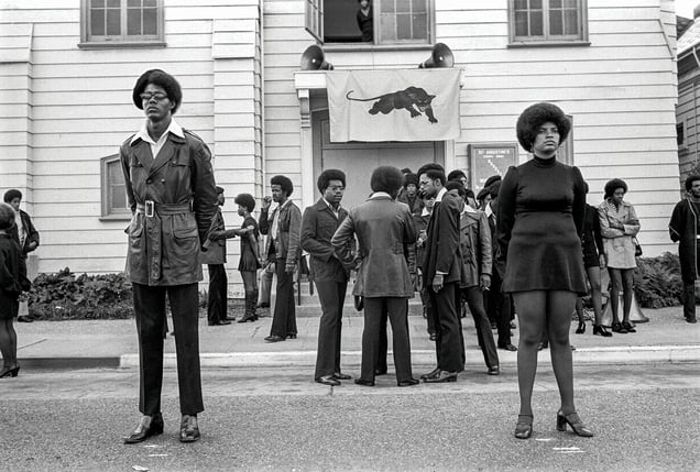 1971, Oakland, California: George Jackson’s funeral at St. Augustine’s Church Glen Wheeler and Claudia Grayson, known as Sister Sheeba, stand in the front. Van Hilliard, known as Van Junior, John Seale and Van Taylor stand near the curb. Clark Bailey, known as Santa Rita, smoking, stands to the left of the sidewalk.