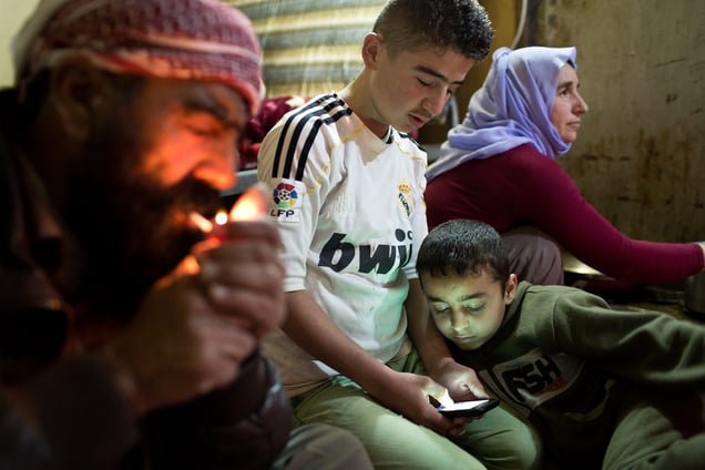 Saïd lights a cigarette while Ezidiar, 9, looks at Serdesht's, 13, phone and Ghazal watches television.The family of Yezidis, displaced from Sinjar, live next to an oil refinery in the Kurdish region of Iraq. The young men run the refinery 24 hours a day with little to no safety equipment. 18/02/15