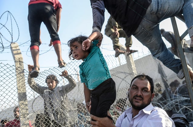 A refugee man holds a young girl as others climb over broken-down border fences to enter Turkish territory; Sanliurfa, Turkey, 14 June 2015.