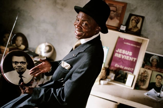 Untitled. The sapeur Severin, also known as the Japanese ambassador, shows a portrait of his father, who was also a sapeur. As one can see in the background, most Congolese are quite religious. Sapeurs tend towards the Catholic or Christian evangelist churches. © Héctor Mediavilla