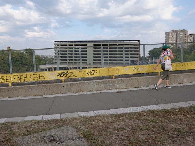 A view toward a multi-storey car park near the railway station in Belgrade, Serbia. The yellow barrier cuts across the frame, separating the pedestrian edge from the large infrastructural volume behind it.