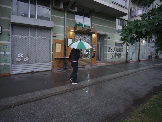 A street in Detelinara, Novi Sad, Serbia. A passerby moves past a ground-floor café embedded in the dense residential fabric of the neighbourhood.