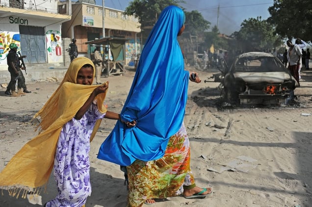 A mother and child walking past the wreckage of a car bomb that killed one person and injured another. Wardhigley District, south of Mogadishu, February 27, 2015. © Mohamed Abdiwahab / AFP