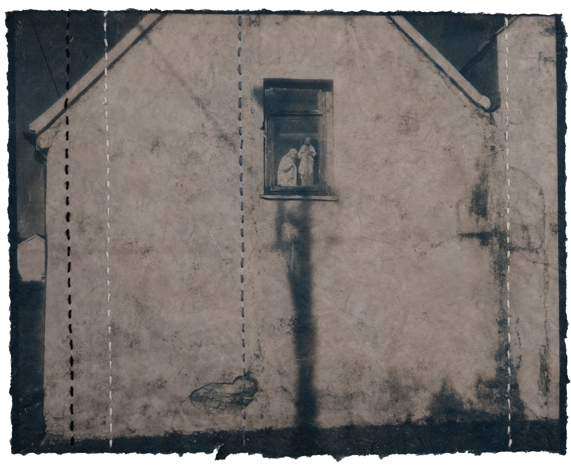 This is an image of a house with a shadow highlighting a window and religious statues. It was made with the cyanotype process onto semi-transparent, hand made paper. It has been toned with black tea and stitched with flax thread. Cyanotype is a process developed in the 1840s, the same time period that Ireland was experiencing a famine. Flax is a material historically used in Ireland for fabric and stitching. The tea tones the brightness of the blue cyanotype to a more subdued brown and blue. The paper is ripped and torn in places and the edges of the paper are deckled. The way the light falls, the window and lines of the shadow emphasize the statues which represent Catholicism, the prevalent religion in the Republic of Ireland. © Patricia Howard